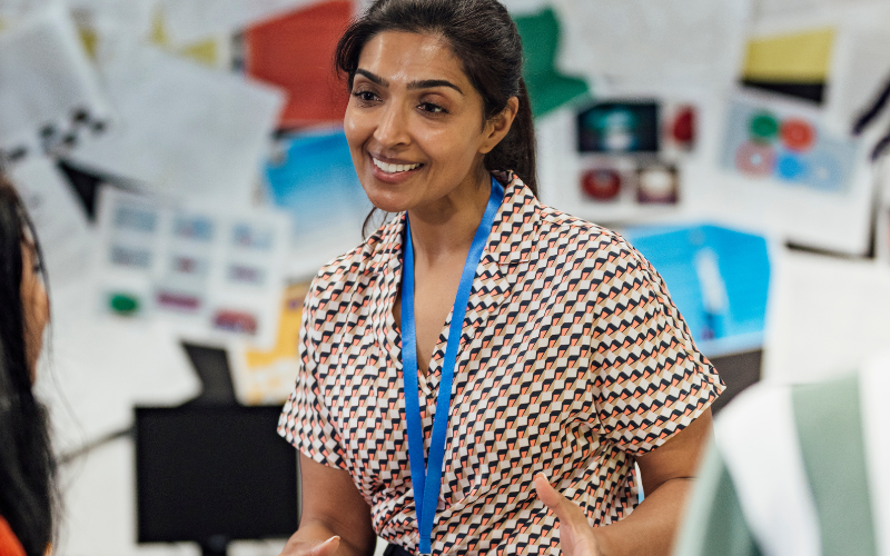 A teacher wearing a patterned blouse and blue lanyard is smiling and talking calmly to another student in a classroom setting.