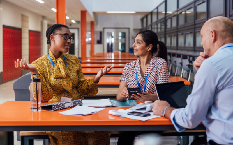 Three teachers are sitting at an orange table in a modern school, engaged in discussion with papers and electronic devices.