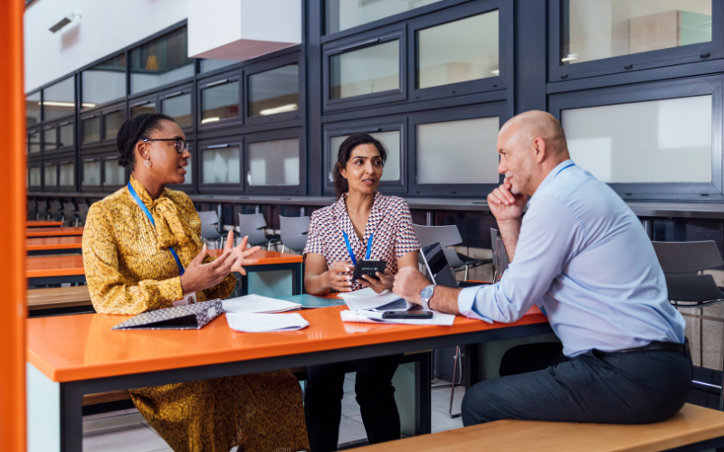 Three teachers are sitting at an orange table in a modern school, engaged in discussion with papers and electronic devices.