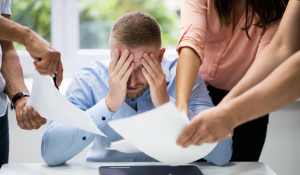 A person sits at a desk holding their head in frustration while surrounded by others handing them papers.