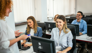 A group of people in a computer lab, with two women seated at a desk and one up front sharing information.
